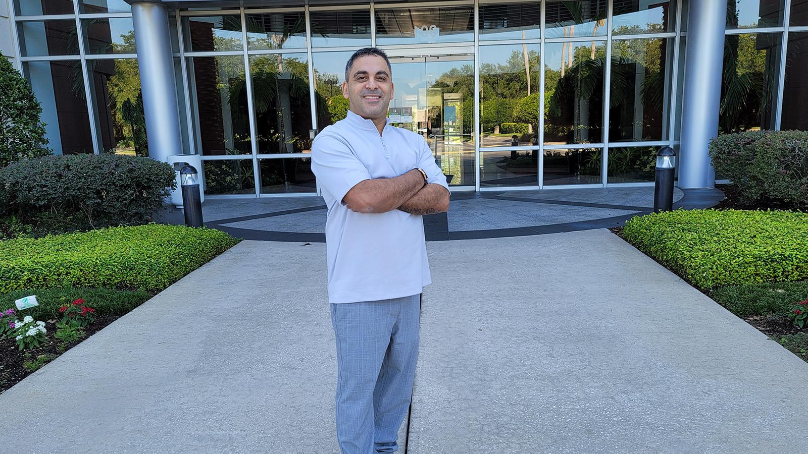 Brian Alvarado双臂交叉站在德勤大楼前微笑着. 他穿着灰色休闲裤和蓝色衬衫.